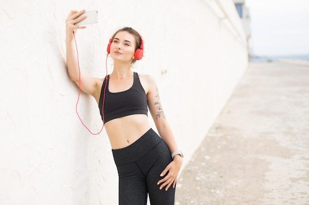 Jovem mulher bonita no top esportivo cinza escuro e leggings com fones de ouvido vermelhos, encostado na parede enquanto sonhadoramente tirando fotos no celular ao ar livre