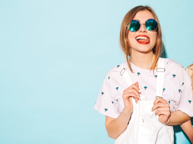 Jovem mulher bonita olhando. Menina na moda em roupas de verão casual amarelo camiseta mostrando a língua em óculos de sol redondos. Fêmea positiva mostra emoções faciais. Modelo engraçado isolado em azul