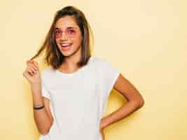 Foto grátis jovem mulher bonita olhando para a câmera. menina na moda casual verão branco camiseta e jeans shorts em óculos de sol redondos. fêmea positiva mostra emoções faciais. modelo engraçado isolado em amarelo