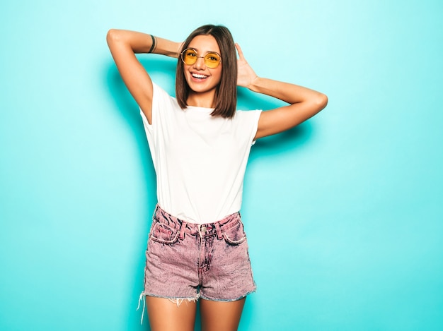 Jovem mulher bonita olhando para a câmera. Menina na moda casual verão branco camiseta e jeans shorts em óculos de sol redondos. Fêmea positiva mostra emoções faciais. Modelo engraçado isolado em azul