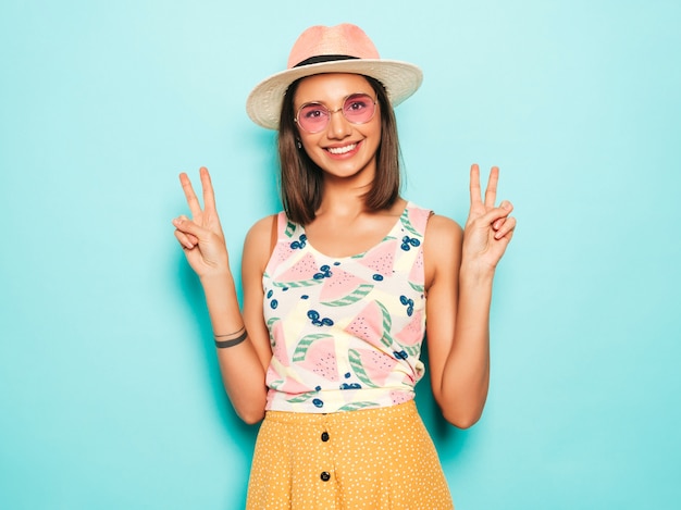 Foto grátis jovem mulher bonita olhando para a câmera no chapéu. menina na moda em camiseta casual verão branco e saia amarela em óculos de sol redondos. fêmea positiva mostra emoções faciais. mostra sinal de paz