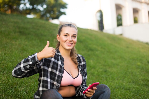 Jovem mulher bonita plus size na camisa casual e top esportivo segurando o celular na mão olhando alegremente na câmera mostrando o polegar para cima gesto enquanto passa o tempo no parque