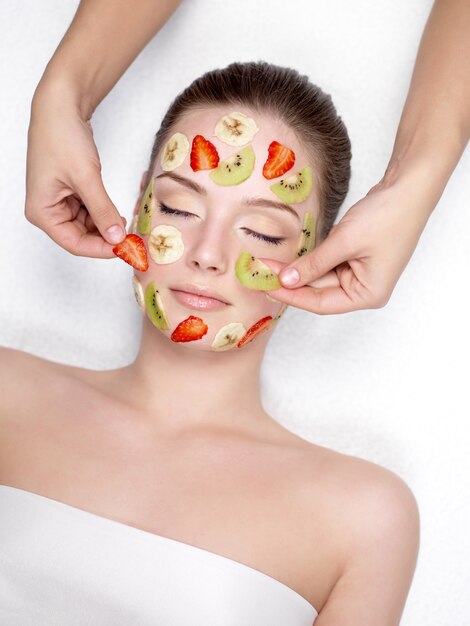 Jovem mulher bonita recebendo máscara facial de frutas cosméticas de morango, banana e kiwi no salão de beleza