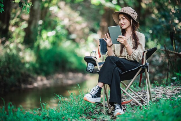 Jovem mulher bonita sentada na cadeira e usar a chamada de vídeo do tablet enquanto acampa no espaço de cópia do parque natural