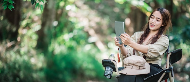 Jovem mulher bonita sentada na cadeira e usar a chamada de vídeo do tablet enquanto acampa no espaço de cópia do parque natural