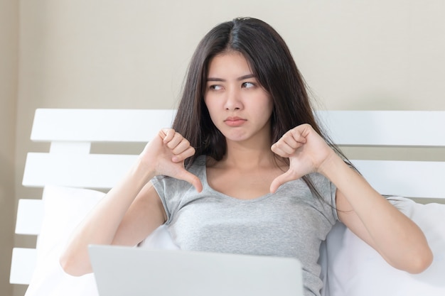 Jovem mulher bonita sentada na cama e usando o computador portátil, ela agindo com thump para baixo se sentir chateada