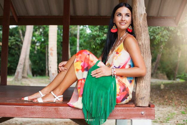 Jovem mulher bonita sexy em um vestido colorido, estilo hippie de verão, férias tropicais, pernas bronzeadas, sandálias, bolsa verde com franja, acessórios, sorrindo, feliz