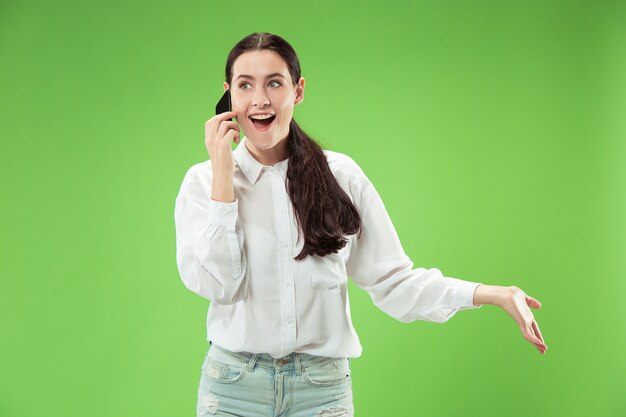 Jovem mulher bonita usando o estúdio do telefone móvel no fundo do estúdio de cor verde. Conceito de emoções faciais humanas. Cores da moda