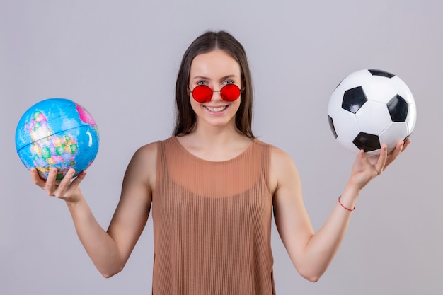 Jovem mulher bonita usando óculos escuros vermelhos segurando bola de futebol e globo com cara feliz sorrindo sobre parede branca