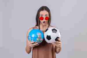 Foto grátis jovem mulher bonita usando óculos escuros vermelhos segurando bola de futebol e globo espantado e surpreso sobre parede branca