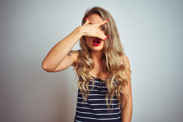 Jovem mulher bonita vestindo camiseta listrada em pé sobre fundo branco isolado espreitando em choque cobrindo o rosto e os olhos com a mão olhando por entre os dedos com expressão envergonhada