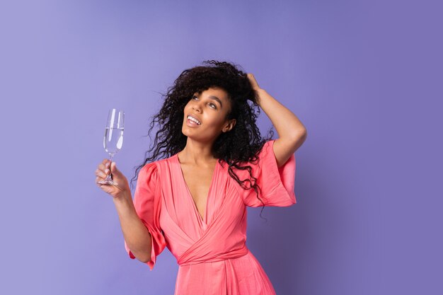 Jovem mulher brasileira feliz com cabelos cacheados em um vestido rosa elegante, posando com uma taça de champanhe sobre a parede roxa. Clima de festa.