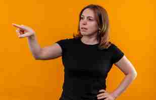 Foto grátis jovem mulher casual apontando e olhando para o lado esquerdo com a mão na cintura em uma parede laranja isolada