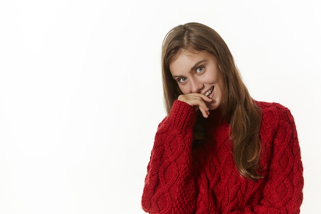 Foto grátis jovem mulher caucasiana brincalhona e sedutora, vestindo um elegante suéter de malha de manga comprida, sorrindo alegremente, segurando a mão nos lábios, posando