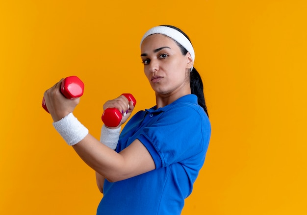 Jovem mulher caucasiana, confiante e esportiva, usando bandana e pulseiras, segurando halteres em laranja com espaço de cópia