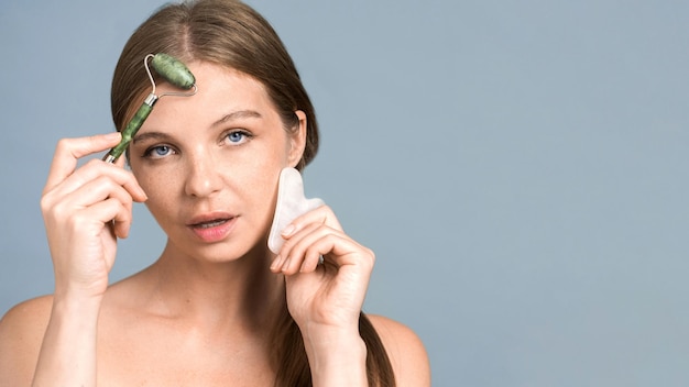 Jovem mulher caucasiana posando com rolo e gua sha para massagem facial olhando para a câmera