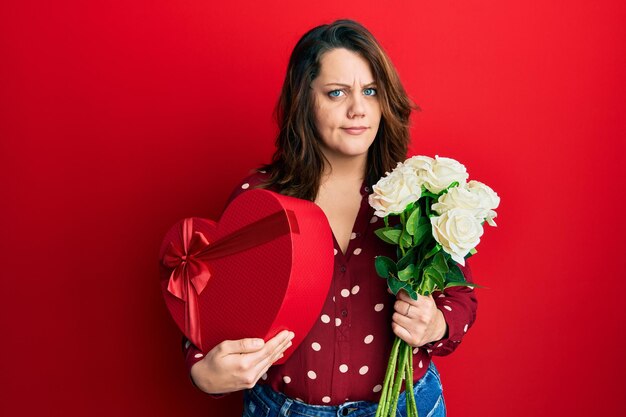 Foto grátis jovem mulher caucasiana segurando presente de dia dos namorados e flores céticas e nervosas franzindo a testa chateadas por causa do problema pessoa negativa