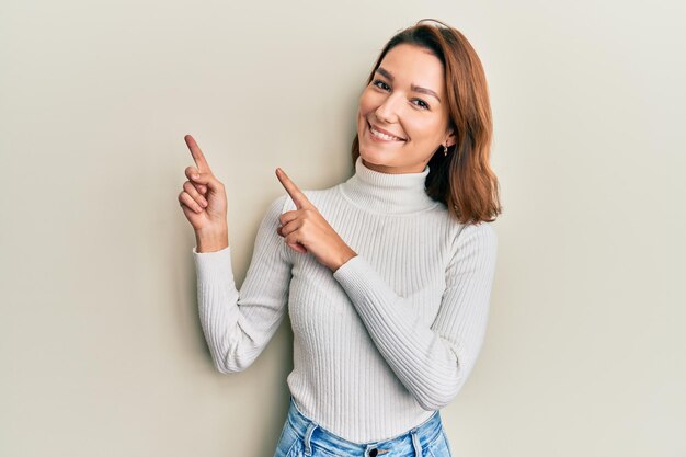 Jovem mulher caucasiana vestindo roupas casuais sorrindo e olhando para a câmera apontando com as duas mãos e os dedos para o lado