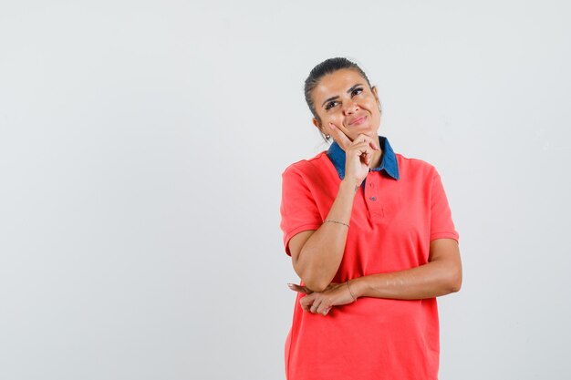 Jovem mulher colocando o dedo indicador na bochecha, pensando em algo em uma camiseta vermelha e olhando pensativa, vista frontal.