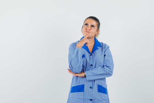 Jovem mulher colocando o dedo indicador no queixo, pensando em gesto em camisa de pijama azul guingão e olhando pensativa. vista frontal.