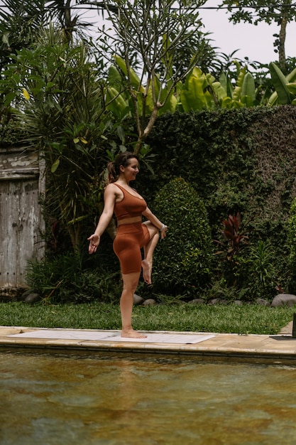 Jovem mulher com aparência positiva do corpo praticando ioga sozinha no deck à beira da piscina na ilha tropical de bali, indonésia. esporte, fitness, conceito de estilo de vida saudável.