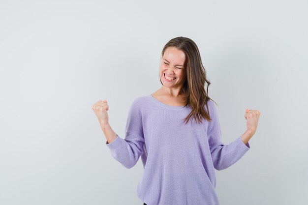 Jovem mulher com blusa lilás mostrando gesto de vencedor e parecendo alegre