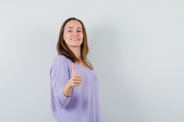 Jovem mulher com blusa lilás mostrando o polegar e parecendo feliz