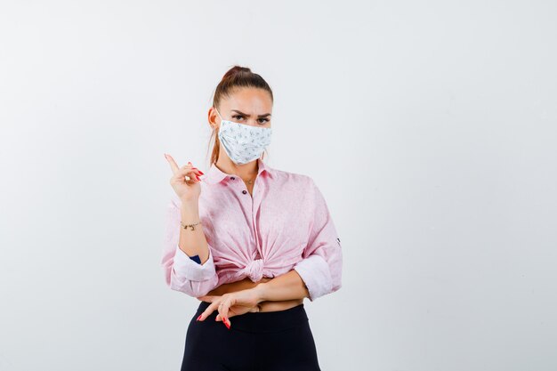 Jovem mulher com camisa, calça, máscara médica, apontando para o canto superior esquerdo e parecendo confiante, vista frontal.