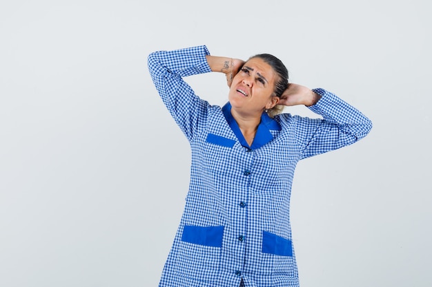 Jovem mulher com camisa de pijama azul guingão, colocando a mão na cabeça e olhando irritada, vista frontal