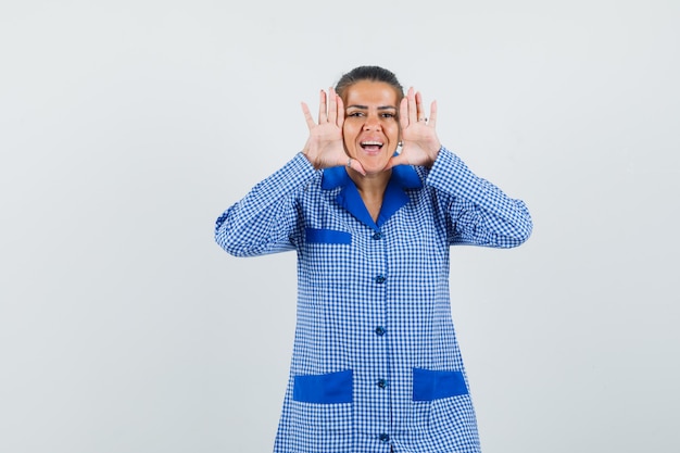 Foto grátis jovem mulher com camisa de pijama azul guingão, colocando a mão perto do rosto, tentando ligar para alguém e olhando feliz, vista frontal.