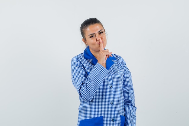 Jovem mulher com camisa de pijama azul guingão colocando o dedo indicador no lábio, mostrando o gesto de silêncio e olhando bonita, vista frontal.