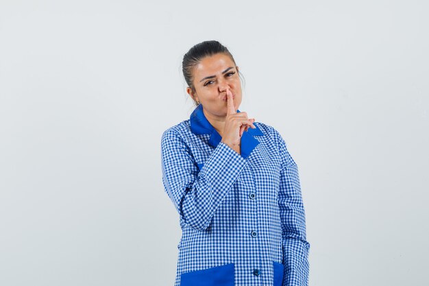 Jovem mulher com camisa de pijama azul guingão colocando o dedo indicador no lábio, mostrando o gesto de silêncio e olhando bonita, vista frontal.