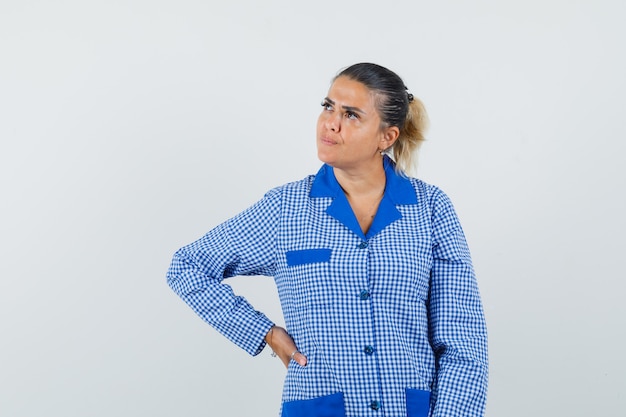 Jovem mulher com camisa de pijama guingão azul, colocando a mão na cintura e olhando pensativa, vista frontal.