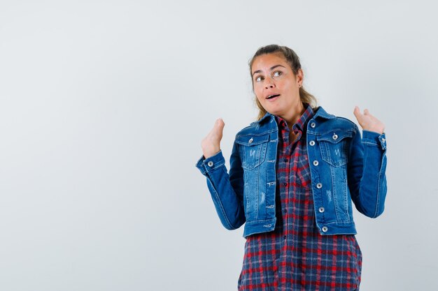 Jovem mulher com camisa, jaqueta, levantando as mãos enquanto olha para cima e parece um sonho, vista frontal.