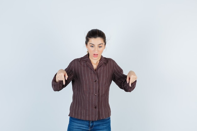 Jovem mulher com camisa listrada, jeans apontando para baixo com o dedo indicador e parecendo surpresa, vista frontal.