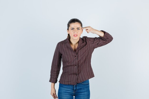 Jovem mulher com camisa listrada, jeans, mostrando o gesto da arma perto da cabeça, fazendo caretas e parecendo aflita, vista frontal.