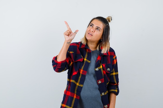 Jovem mulher com camisa quadriculada apontando para o canto superior esquerdo e parecendo confiante