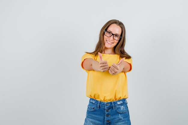 Jovem mulher com camiseta, shorts e óculos mostrando os polegares para cima e sorrindo