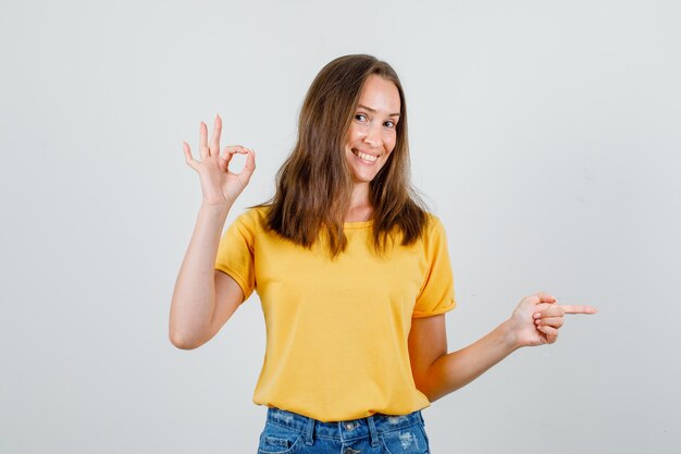 Jovem mulher com camiseta, shorts mostrando sinal de ok enquanto aponta para longe e parece alegre