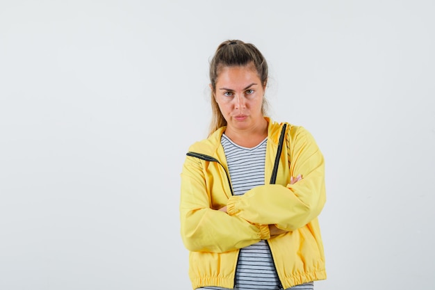 Jovem mulher com casaco, t-shirt em pé com os braços cruzados e olhando sério, vista frontal.