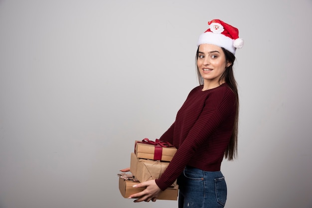 Jovem mulher com chapéu de Papai Noel com presentes de Natal em fundo cinza.