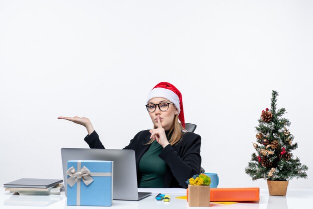 Jovem mulher com chapéu de Papai Noel e óculos, sentada à mesa com uma árvore de natal e um presente