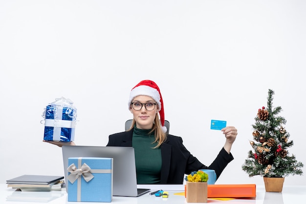 Jovem mulher com chapéu de papai noel e óculos, sentada à mesa segurando um presente de natal e um cartão do banco