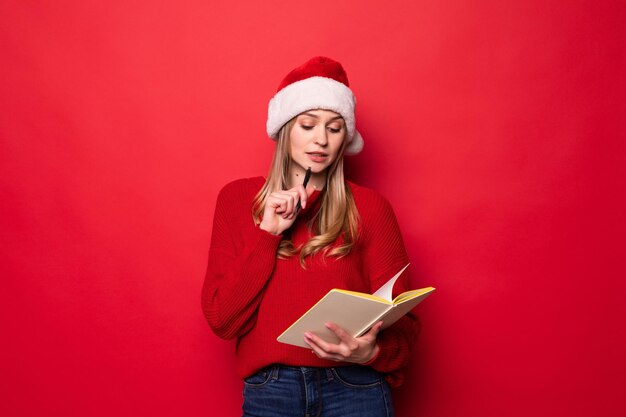 Jovem mulher com chapéu de Papai Noel segura um bloco de notas nas mãos e morde uma caneta, verificando a lista de presentes ou crianças com bom comportamento isoladas em uma parede vermelha.,