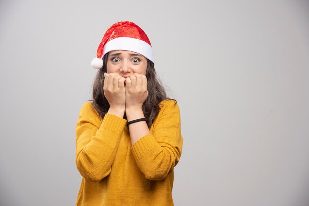 Jovem mulher com chapéu de Papai Noel vermelho posando sobre uma parede branca.