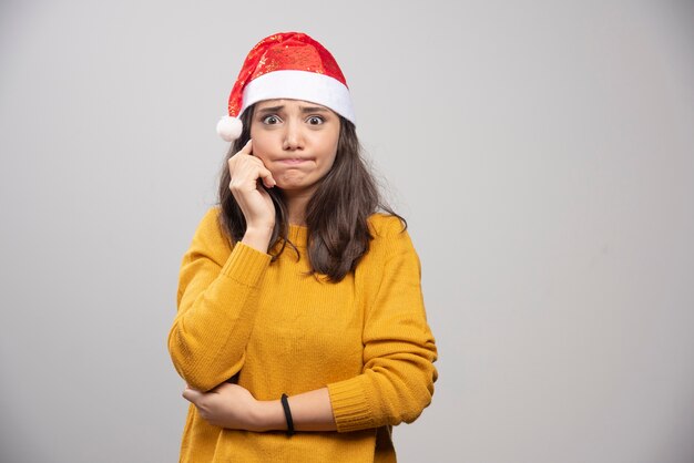 Jovem mulher com chapéu de Papai Noel vermelho posando sobre uma parede branca.