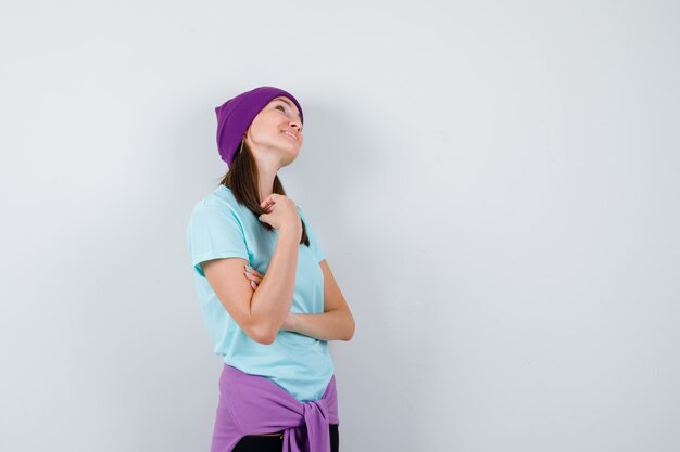 Jovem mulher com fios de cabelo, olhando para cima em t-shirt azul, gorro roxo e olhando alegre, vista frontal.