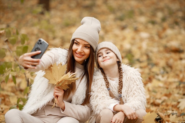 Jovem mulher com garotinha faz uma selfie na floresta de outono. Morena sentada perto de sua filha. Menina vestindo suéter bege e mãe vestindo roupas brancas.