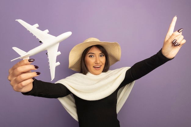 Jovem mulher com lenço branco no chapéu de verão mostrando o avião de brinquedo feliz e alegre sorrindo apontando com o dedo indicador para cima