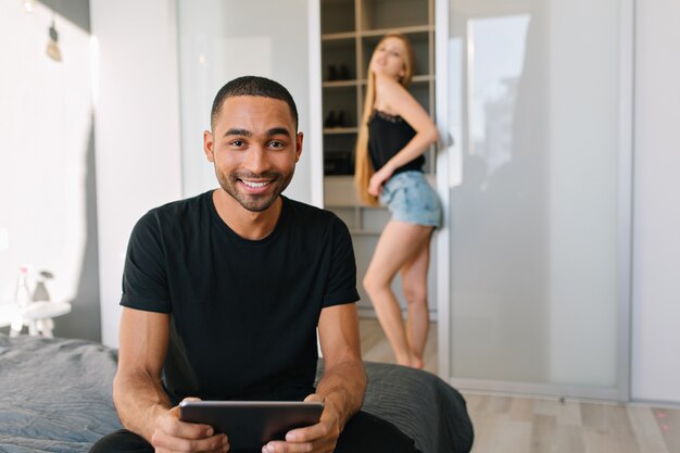 jovem mulher com longos cabelos loiros em shorts, atrás de um cara alegre bonito na cama, sorrindo com o tablet. Apartamento moderno, casal adorável, se divertindo, verdadeiras emoções